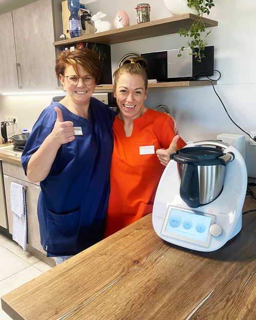 Vorwerk: Foto: Zwei Frauen mittleren Alters stehen eng beieinander in einer Küche, lächeln und bilden mit jeweils einer Hand zusammen das Herz-Zeichen. Zwei Freundinnen. Neben ihnen thront der brandneue Thermomix® TM7.
