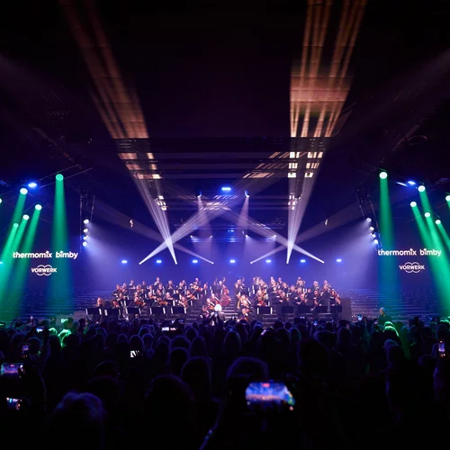 Vorwerk: Foto: Im vierten Foto ist die ganze Halle mit den Spots des Laserlichts durchzogen. Auf der Bühne ist ein großes klassisches Orchester angestrahlt, das musiziert.