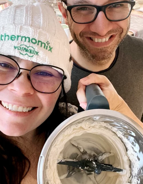 Vorwerk: Foto: Ein fröhliches Paar hält lachend die Schüssel voller Teig eines Thermomix® in die Kamera. Die Frau trägt eine Mütze mit Thermomix® Logo. Beide sehen sehr glücklich aus.