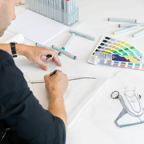 Vorwerk: Foto: Ein Mann ist mit einem Designprojekt beschäftigt und arbeitet konzentriert an einem Schreibtisch, der mit Designwerkzeugen und digitalen Geräten gefüllt ist.