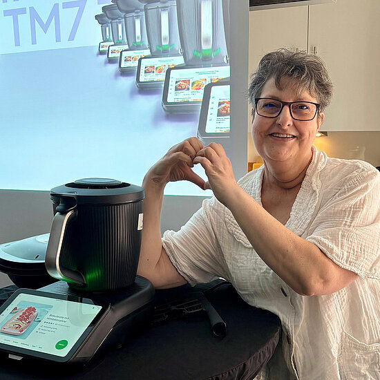 Vorwerk: Foto: Eine ältere Frau formt ein Herz mit Händen. Neben ihr ist ein TM7 zu sehen.
